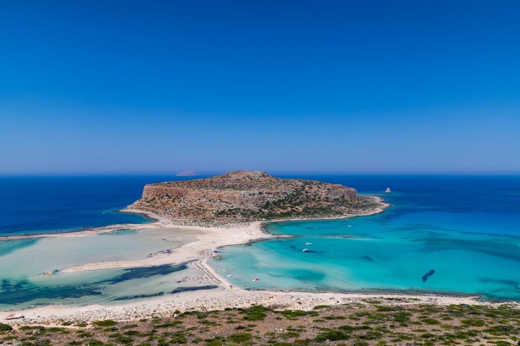 Laguna Balos z turkusową wodą i mielizną prowadzącą do wyspy, widziana z punktu widokowego na wzgórzu.