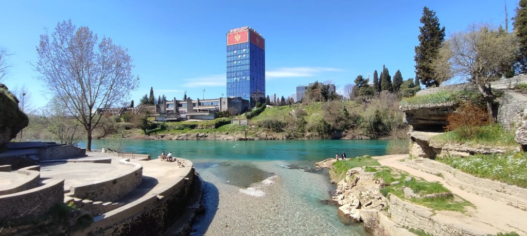 Fragment niebieskiej rzeczki otoczony zieloną roślinnością, z widokiem na mały niebieski szklany wieżowiec z flagą Czarnogóry w tle, w Podgoricy.