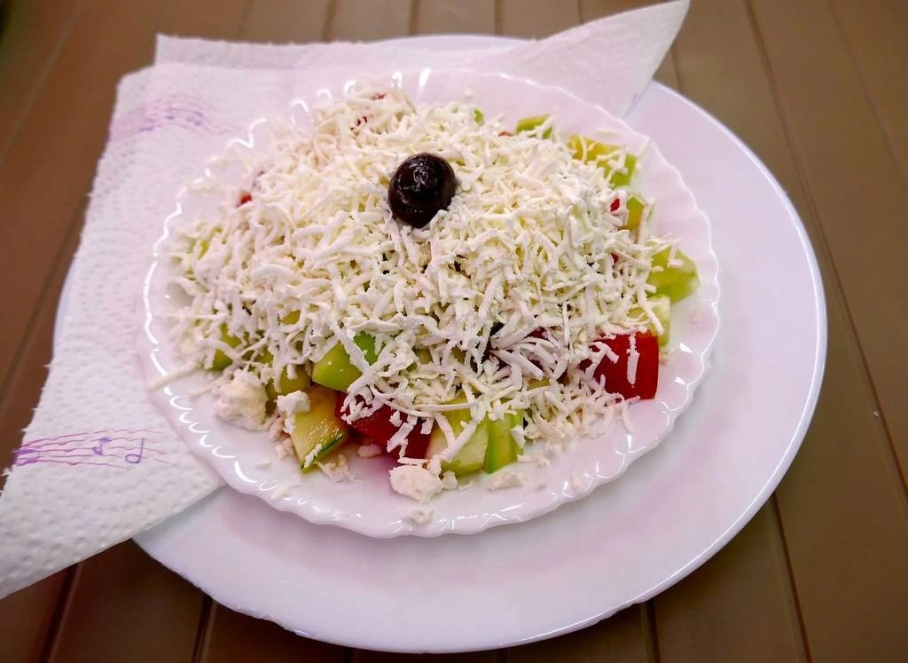 Sałatka shopska z warzywami, przykryta serem owczym i udekorowana czarną oliwką na środku.