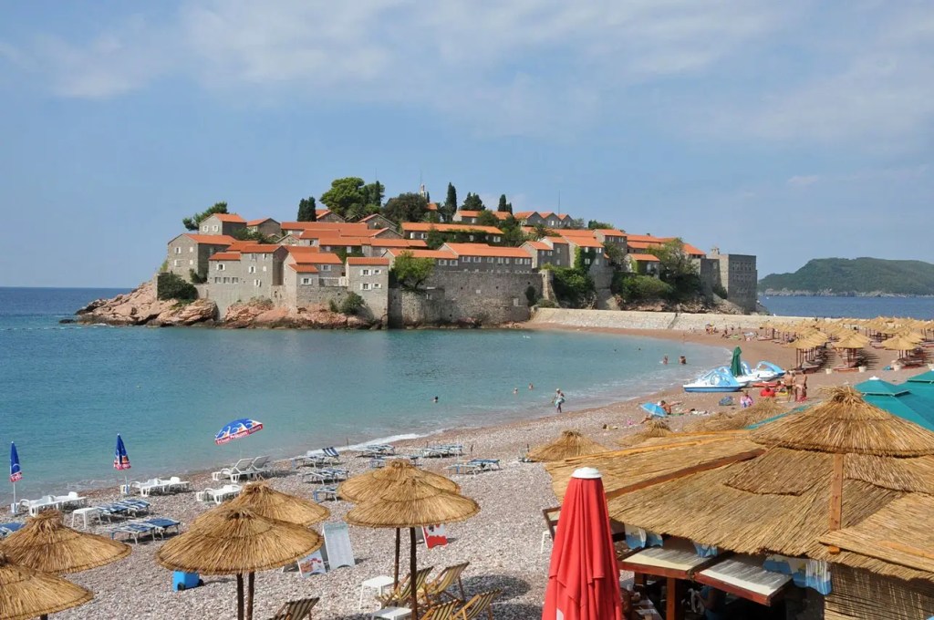 Sveti Stefan widziany z pobliskiej plaży w ciągu dnia, z charakterystycznym półwyspem i budynkami otoczonymi morzem.