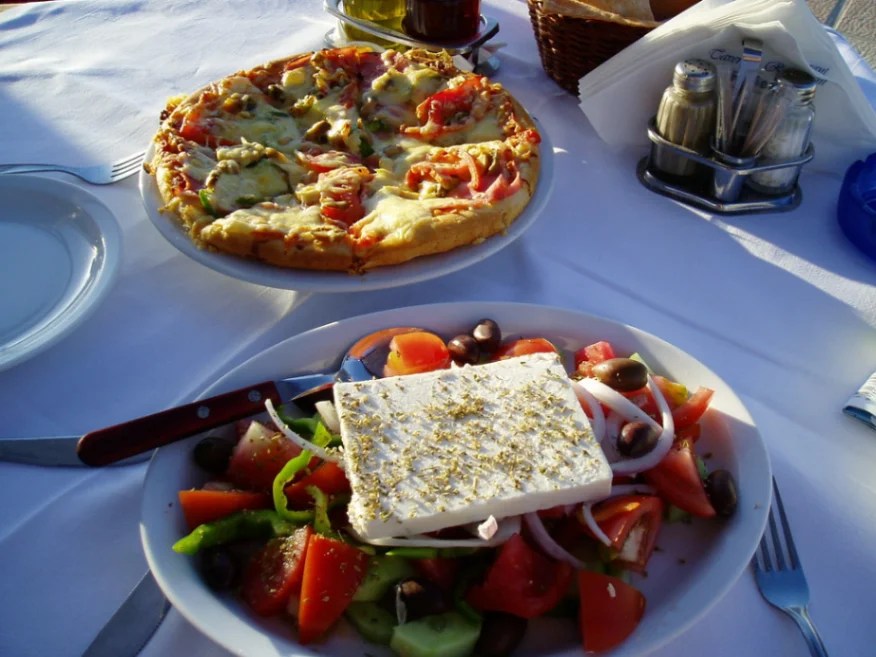 Stół z tradycyjną kreteńską sałatką z dużym kawałkiem fety posypanej oregano i małą pizzą na osobnym talerzu.