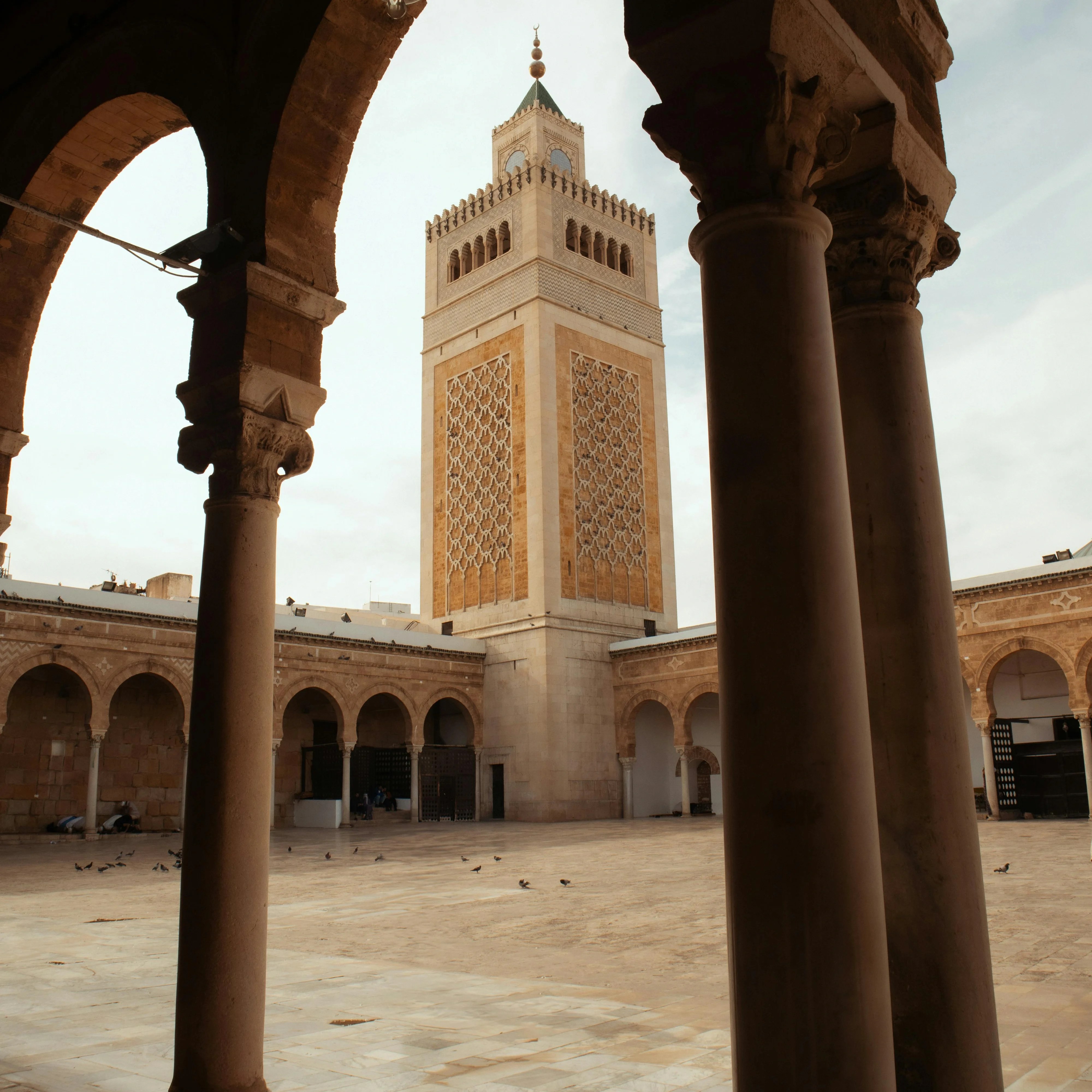 Widok na główną wieżę Meczetu Zaytuna z placu otoczonego kolumnami i zadaszeniem w medynie Tunisu.